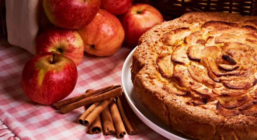 Torta mele, noci e cannella dorata, con mele fresche e bastoncini di cannella su una tovaglia a quadri rosa.