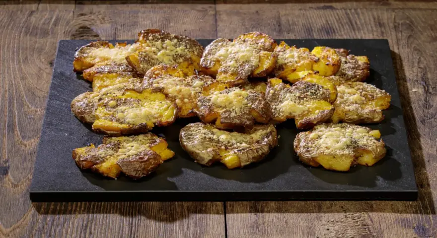 Patate schiacciate al forno, dorate e croccanti, cosparse di formaggio grattugiato e spezie, servite su una pietra scura.
