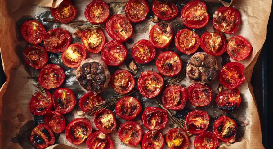 Pomodori al forno, caramellati e succosi, con aglio arrostito e rosmarino su carta forno.