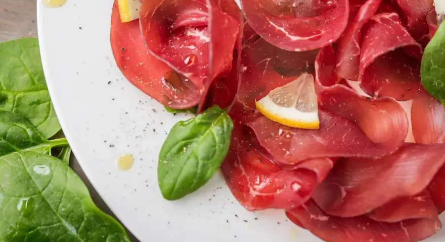 Fette sottili di bresaola di manzo su un piatto bianco, guarnite con spinaci freschi, spicchi di limone e un filo d'olio.