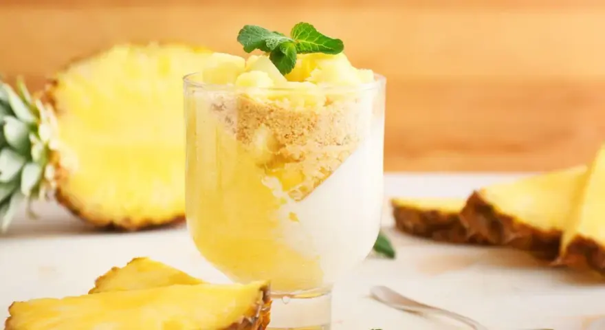 Delizioso dolce con ananas fresco, yogurt e crumble. Un bicchiere pieno di gusto tropicale, con fette di ananas e menta a guarnire.