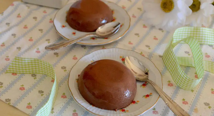 Due budini al cioccolato con amido di mais, serviti su piattini floreali con cucchiaini d'argento. Nastro a quadretti verdi completa la scena.