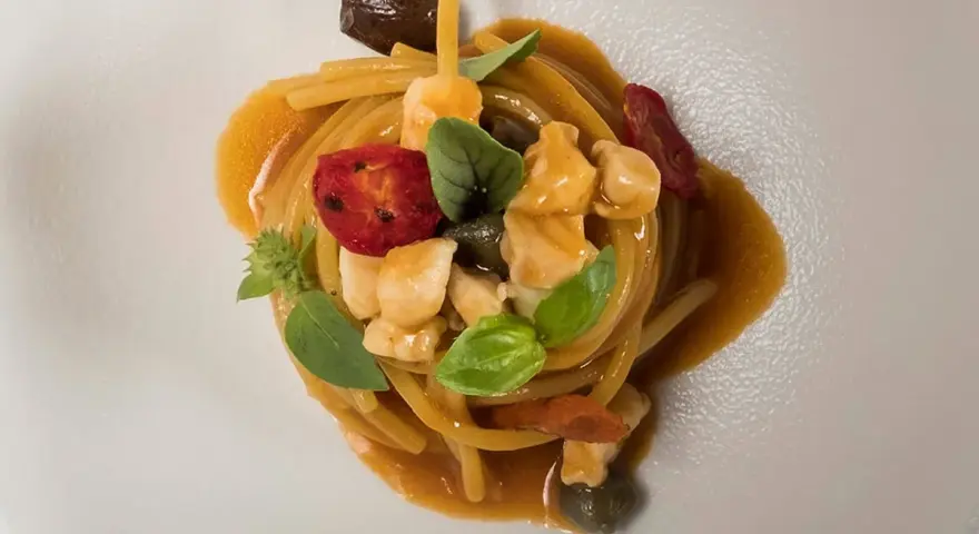 Un piatto di pasta con il pesce, condita con pomodorini, olive, capperi e basilico, servita su un piatto bianco.