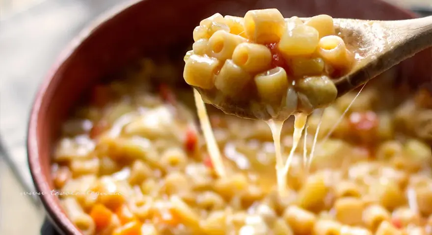 Un cucchiaio solleva una porzione fumante di pasta e patate veloce, filante di formaggio fuso, in una ciotola di terracotta.