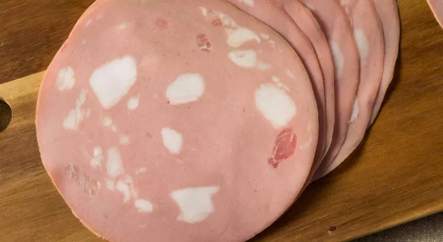 Fette di mortadella su tagliere di legno. Scopri come si fa la mortadella, un salume rosa con cubetti di grasso bianco.