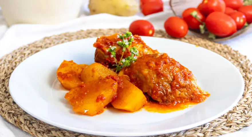 Piatto di cosciotto di tacchino in umido con patate, guarnito con erbe aromatiche. Sullo sfondo, pomodori e patate fresche.