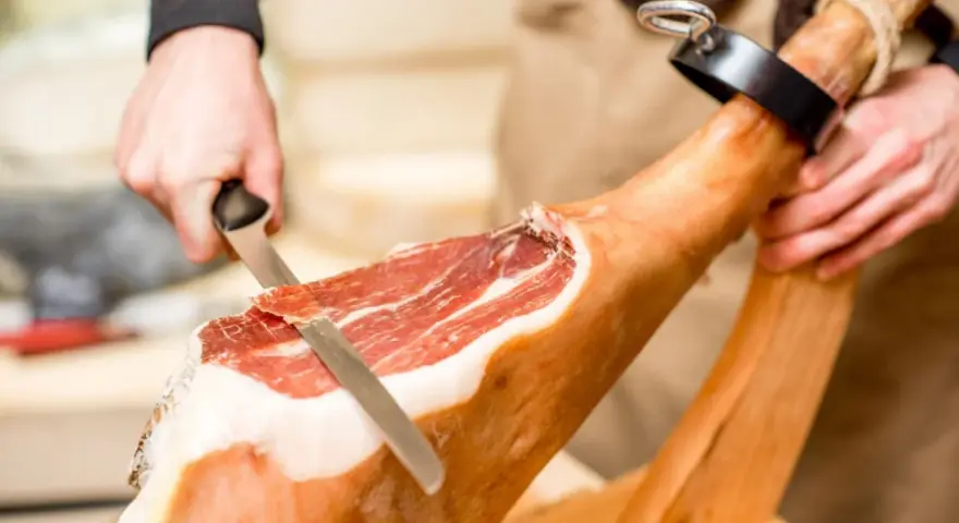 Mani esperte dimostrano come tagliare il prosciutto crudo con un coltello affilato, creando fette sottili e gustose.