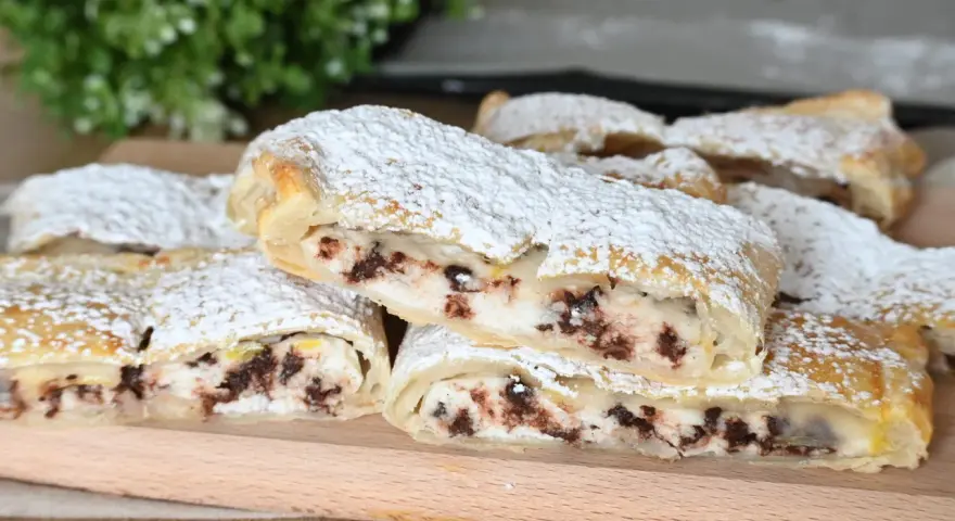 Rotolo di sfoglia con ricotta dolce e gocce di cioccolato, spolverato di zucchero a velo, su un tagliere di legno.