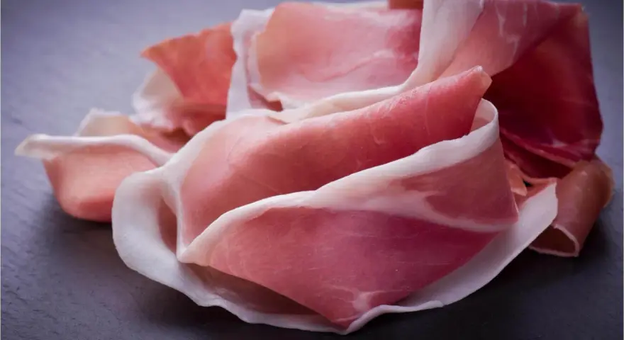 Fette di prosciutto crudo, un insaccato dal colore rosato e venature bianche, disposte su una superficie scura.
