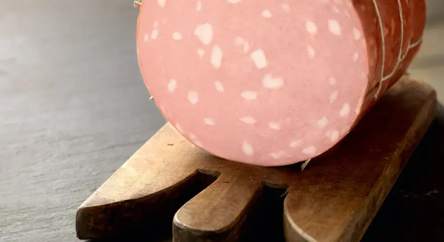 Una fetta di mortadella è cotta, con i suoi tipici cubetti bianchi di grasso, posata su un tagliere di legno.