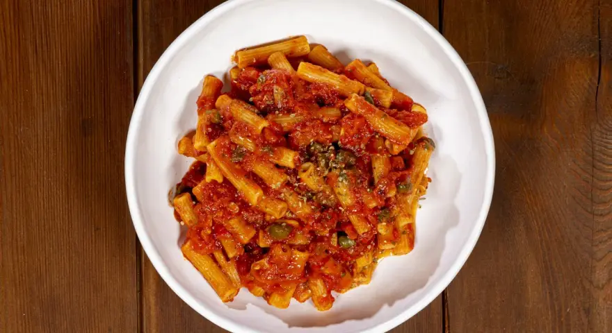 Un piatto di pasta alla pizzaiola, con rigatoni conditi con sugo di pomodoro, capperi e origano, servito in una ciotola bianca su un tavolo di legno scuro.