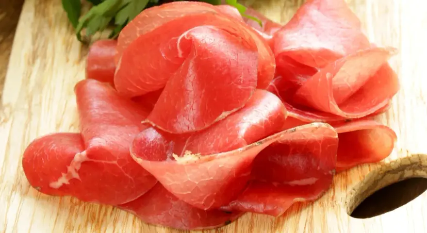 Fettine sottili di salume simile alla bresaola, rosse e lucide, disposte su un tagliere di legno con un ciuffo di prezzemolo.