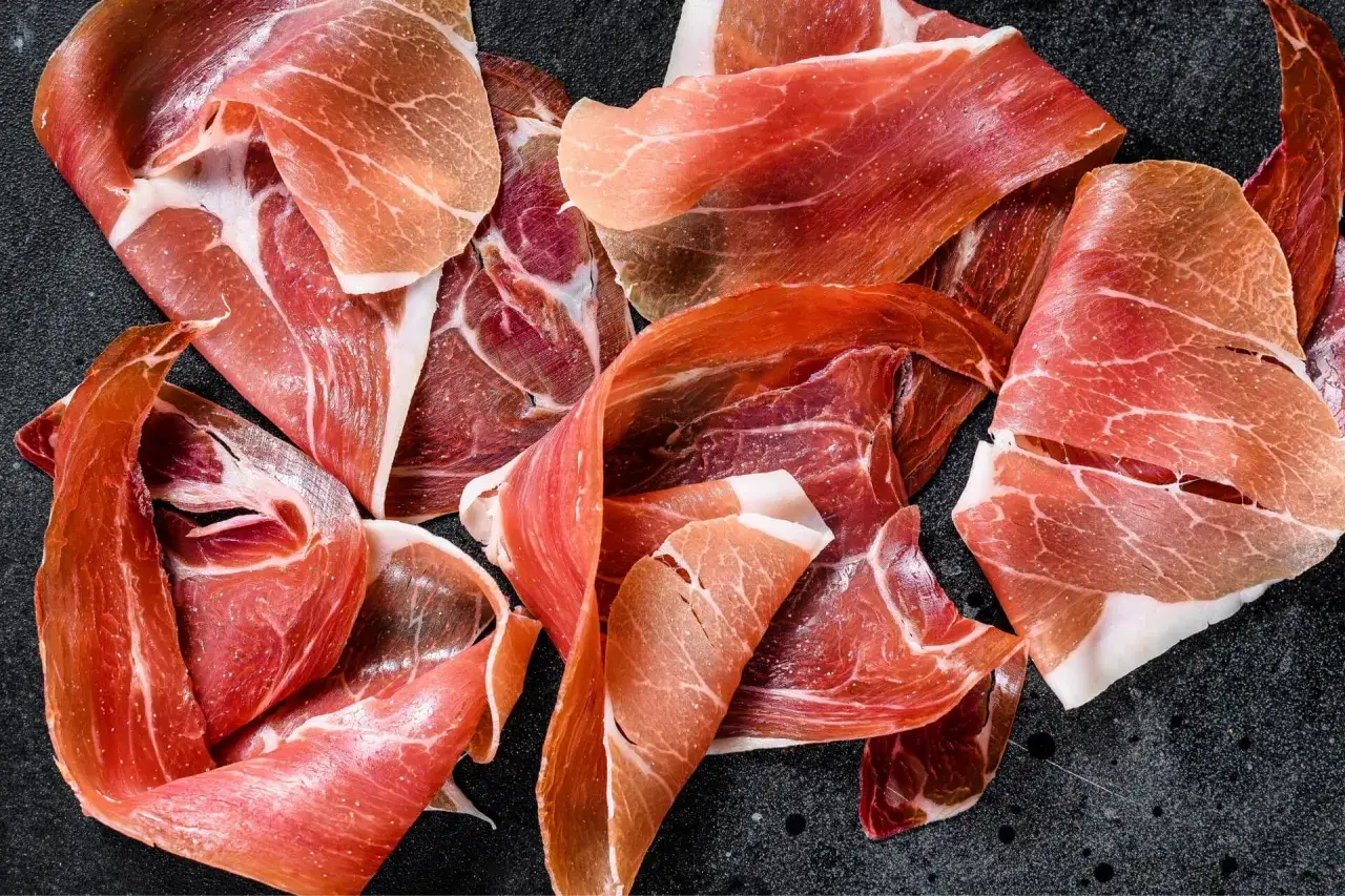 Fette sottili di prosciutto crudo, un insaccato dal colore rosato e venature bianche, disposte su una superficie scura.