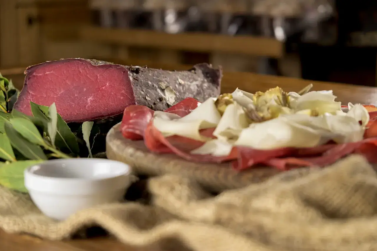 Un pezzo di salume simile alla bresaola, affettati misti e verdure su un tagliere di legno.