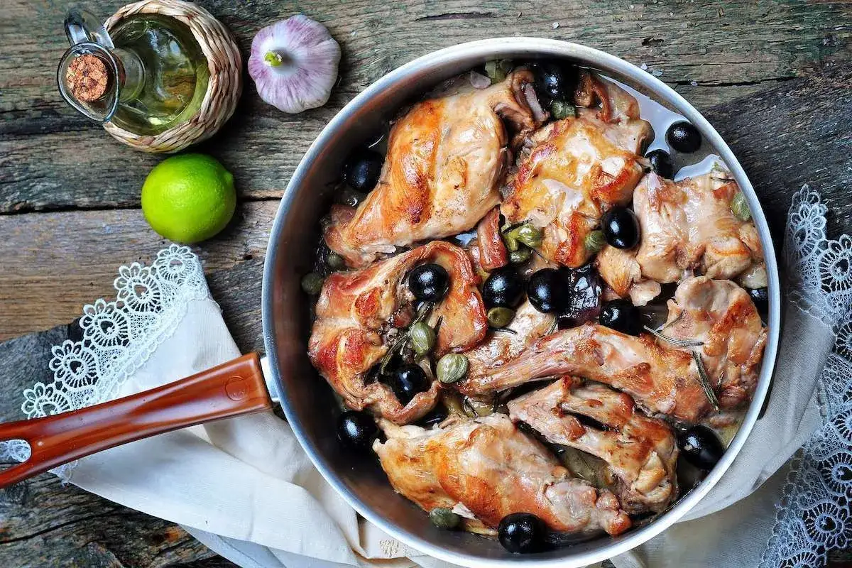 Un saporito coniglio alla ligure in padella, con olive nere, capperi e rosmarino. Ingredienti freschi come aglio, lime e olio d'oliva completano la scena.