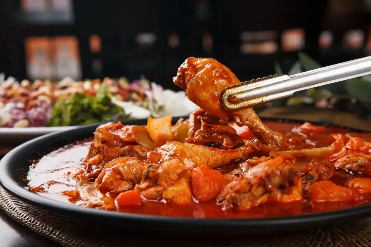 Un succulento fricc&ograve; di pollo in una salsa rossa piccante, con pezzi di verdura. Una pinza solleva un pezzo di pollo.