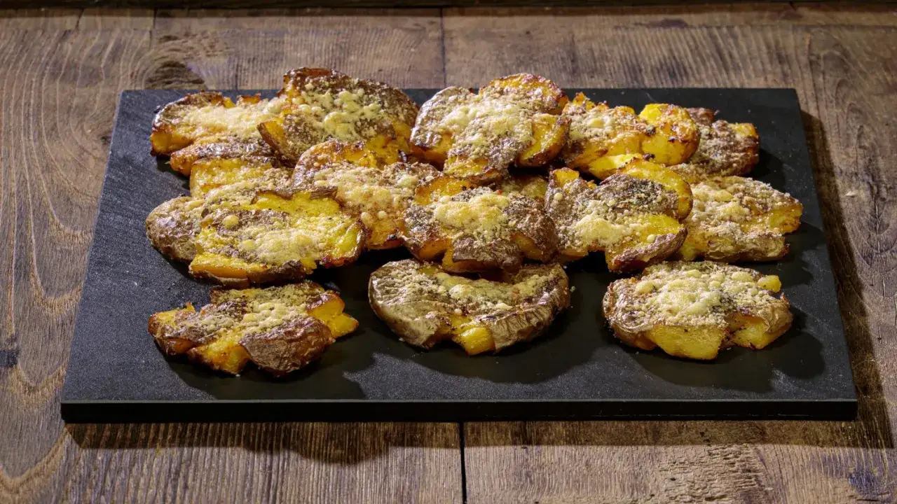 Patate schiacciate al forno, dorate e croccanti, cosparse di formaggio grattugiato e spezie, servite su una pietra scura.