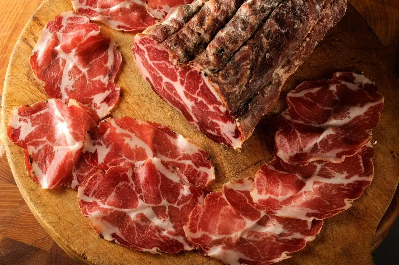 Fette di coppa su tagliere di legno, con un pezzo intero di coppa stagionata. Un assaggio di cos'&egrave; la coppa, un salume pregiato.
