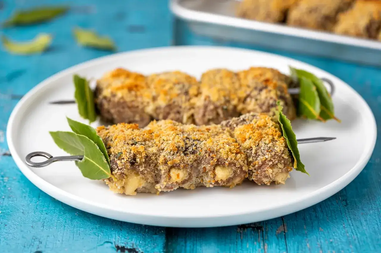 Involtini alla messinese su spiedini, impanati e dorati, guarniti con foglie di alloro su un piatto bianco.