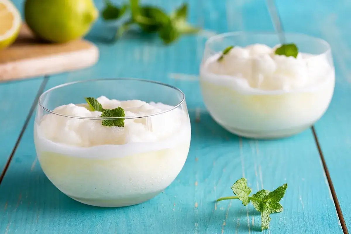 Sorbetto al limone senza gelatiera: due coppe di fresco sorbetto, guarnite con menta, su un tavolo di legno blu.