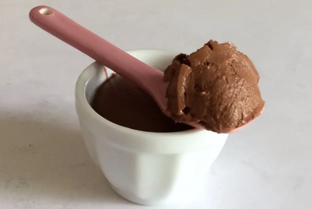 Un cucchiaio rosa raccoglie una porzione di cremoso budino al cioccolato con amido di mais da una ciotolina bianca.