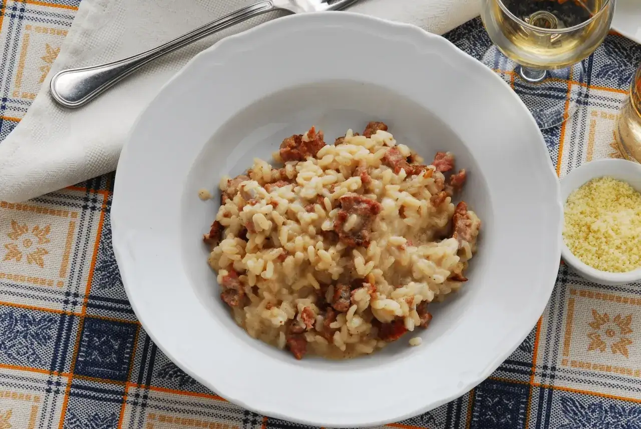 Un piatto di risotto alla pilota, cremoso e ricco, con pezzetti di salsiccia. Accanto, una ciotolina di formaggio grattugiato e un bicchiere di vino bianco.