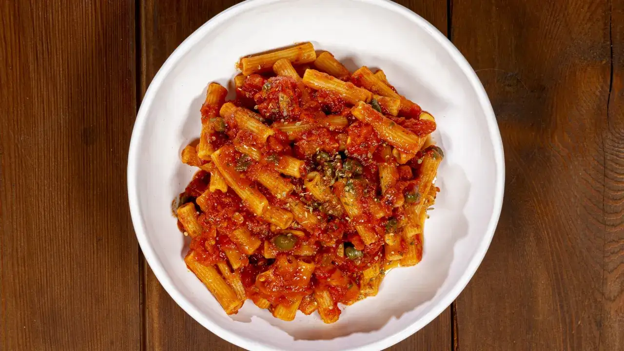 Un piatto di pasta alla pizzaiola fumante, con rigatoni conditi con sugo rosso, capperi e origano, servito in una ciotola bianca su un tavolo di legno scuro.