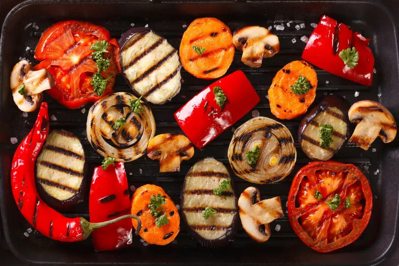 Varietà di contorni per grigliata: pomodori, melanzane, cipolle, peperoni, carote e funghi, cotti su una piastra.