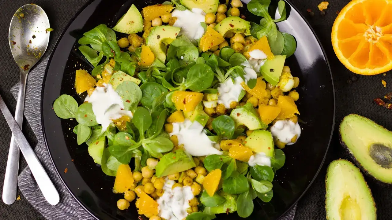 Deliziosa insalata di legumi con ceci, avocado, arancia e un condimento cremoso, guarnita con erbe fresche.