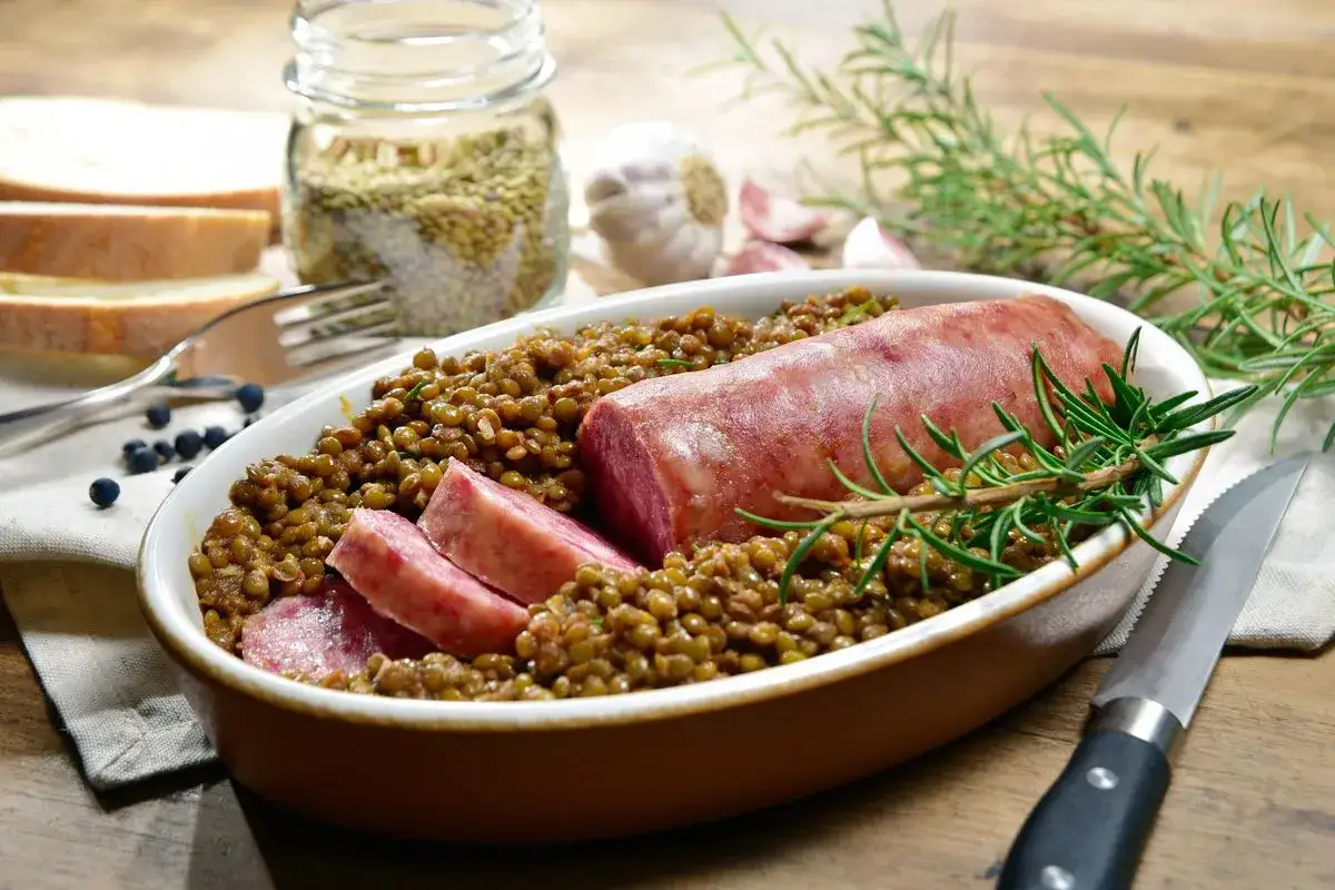 Piatto di lenticchie con cotechino affettato, un classico su come si fa il cotechino. Rosmarino, aglio e pane completano la scena.