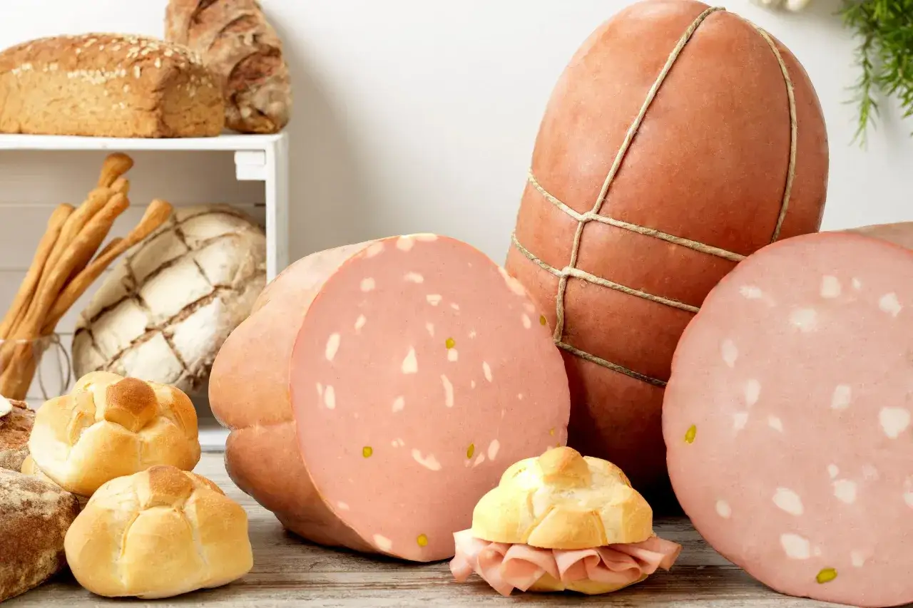 La mortadella &egrave; cotta, con fette rosa e bianche, accanto a pane fresco e grissini.