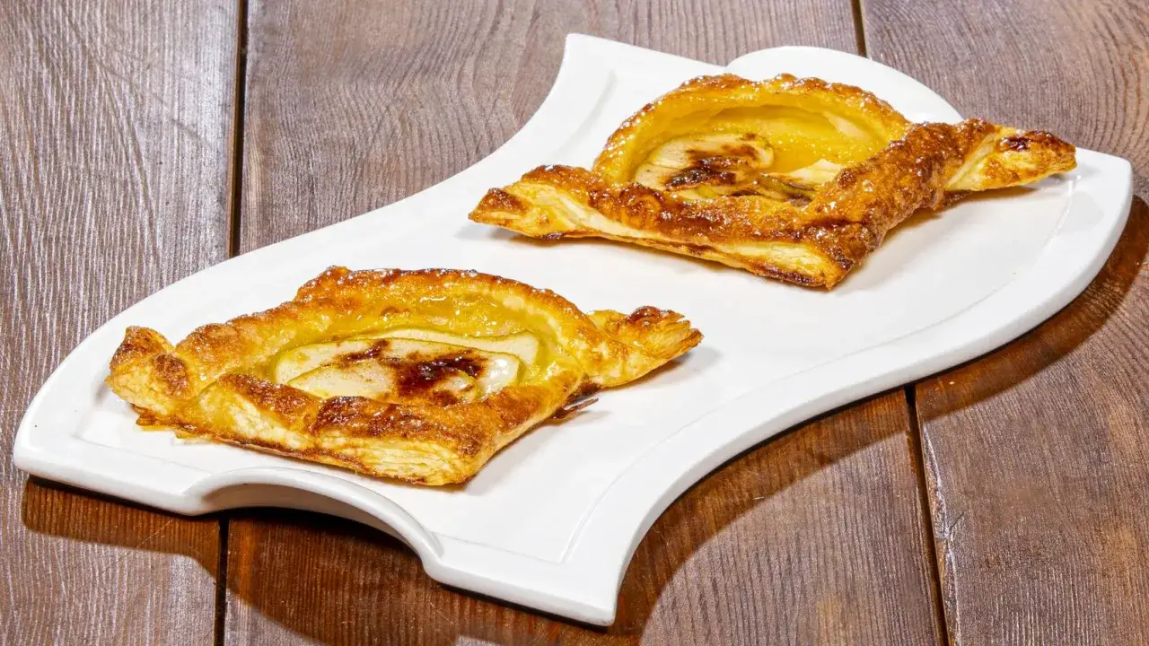 Due sfogliatine di mele dorate e lucide, con mele caramellate e un pizzico di zucchero a velo, su un piatto bianco.