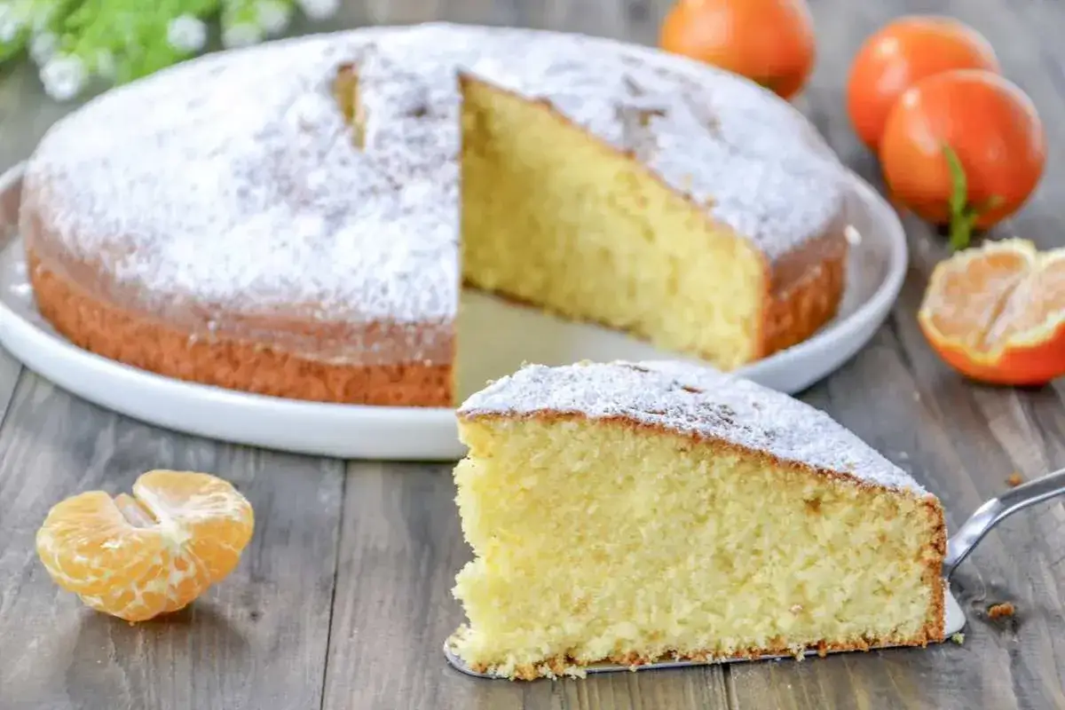 Una soffice torta di clementine, cosparsa di zucchero a velo, con una fetta servita su una paletta. Arance fresche completano la scena.