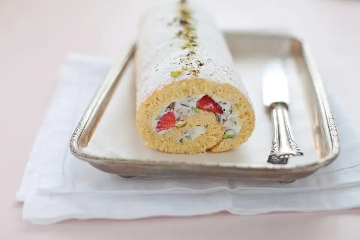 Rotolo di sfoglia con ricotta dolce, fragole e pistacchi, spolverato di zucchero a velo, su un vassoio d'argento.