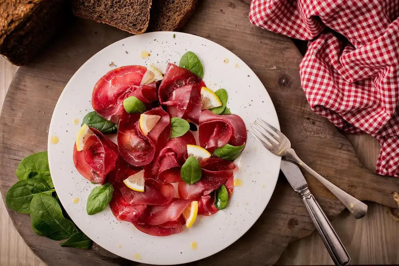 Piatto di bresaola di manzo affettata finemente, guarnita con foglie di spinacino fresco e spicchi di limone. Accanto, pane scuro e tovagliolo a quadri rossi.
