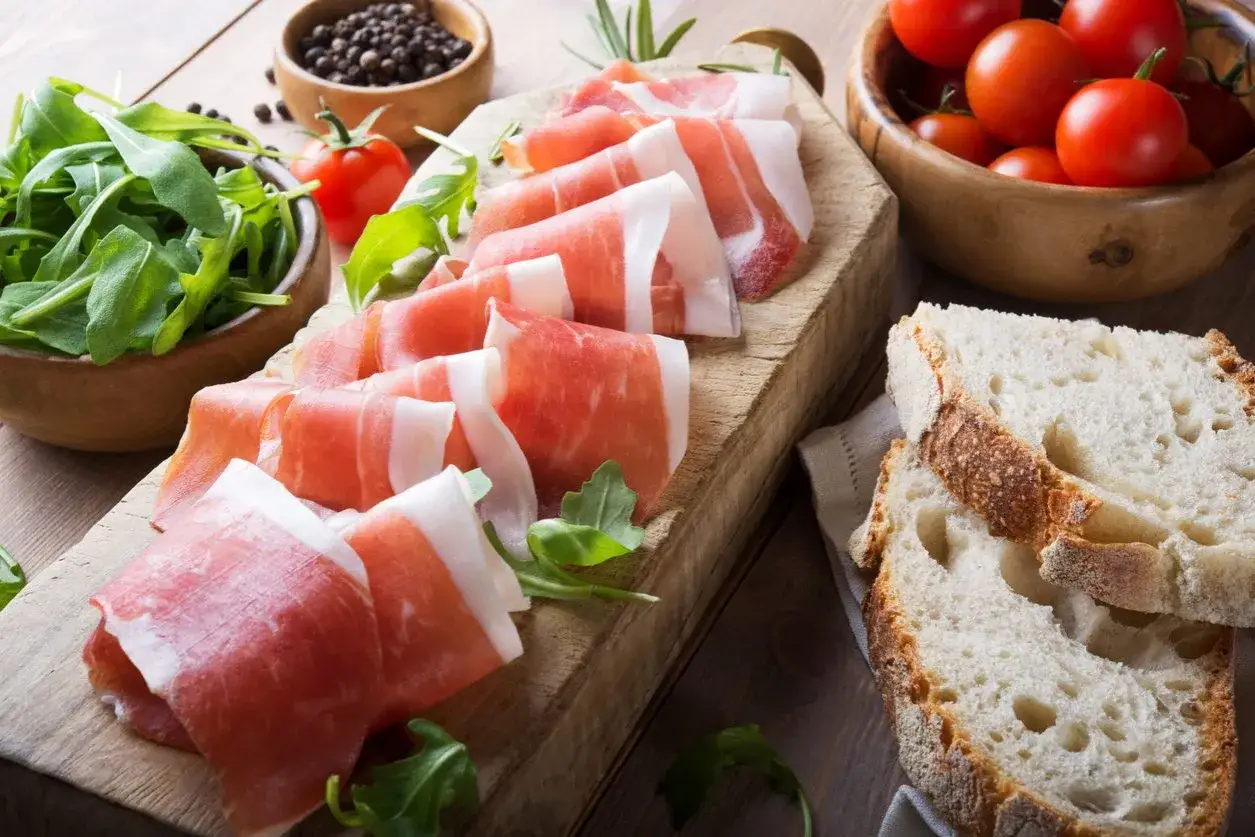 Fette di prosciutto crudo su tagliere con rucola, pomodorini e pane. Scopri come utilizzare il grasso del prosciutto crudo per insaporire i tuoi piatti.