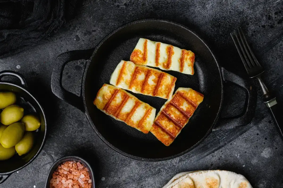 Quattro fette di formaggio da fare alla piastra, dorate e con striature invitanti, in una padella di ghisa. Accanto, olive verdi e sale rosa.