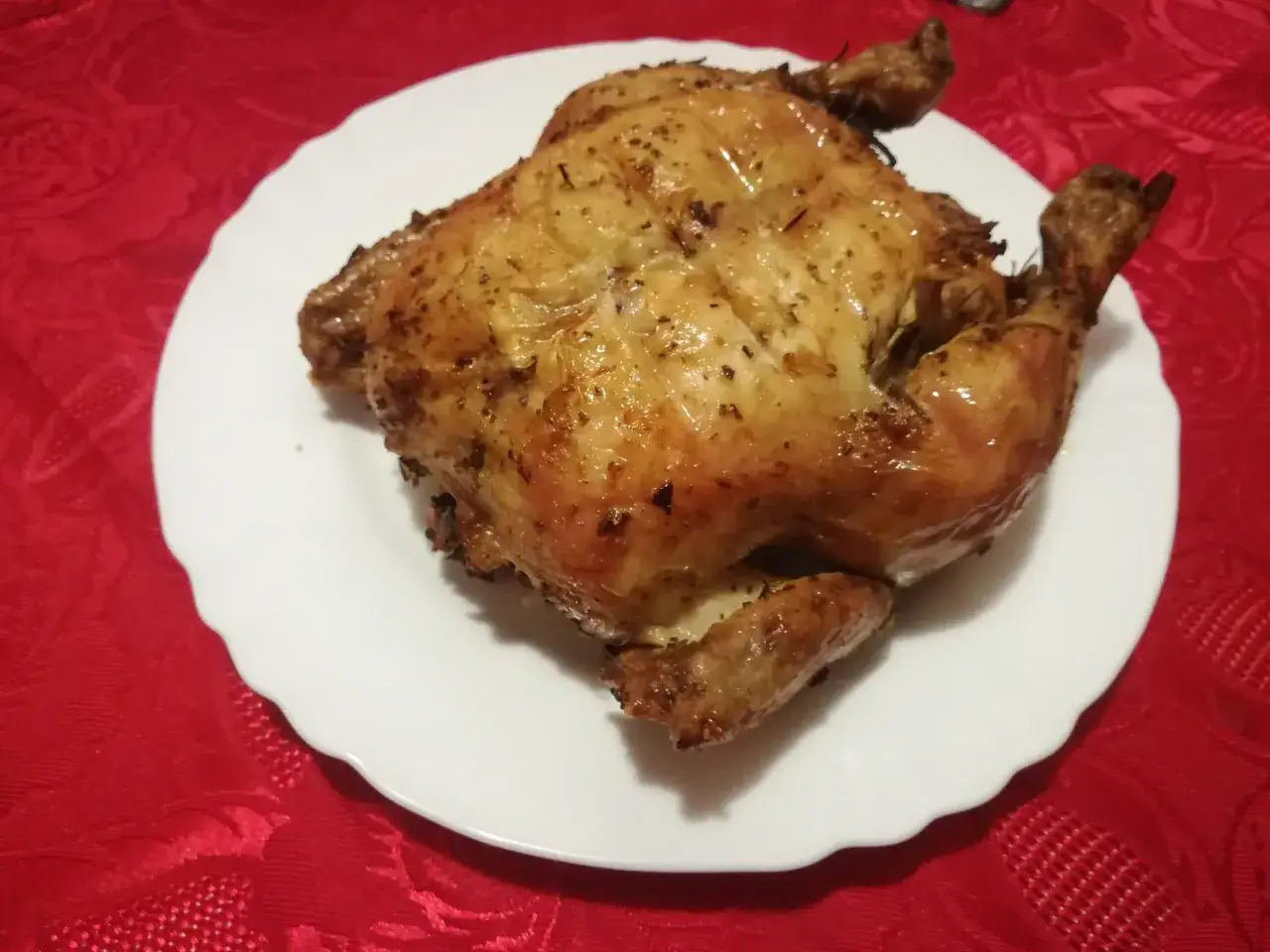 Un succulento pollo allo spiedo dorato, condito con erbe aromatiche, servito su un piatto bianco su una tovaglia rossa.