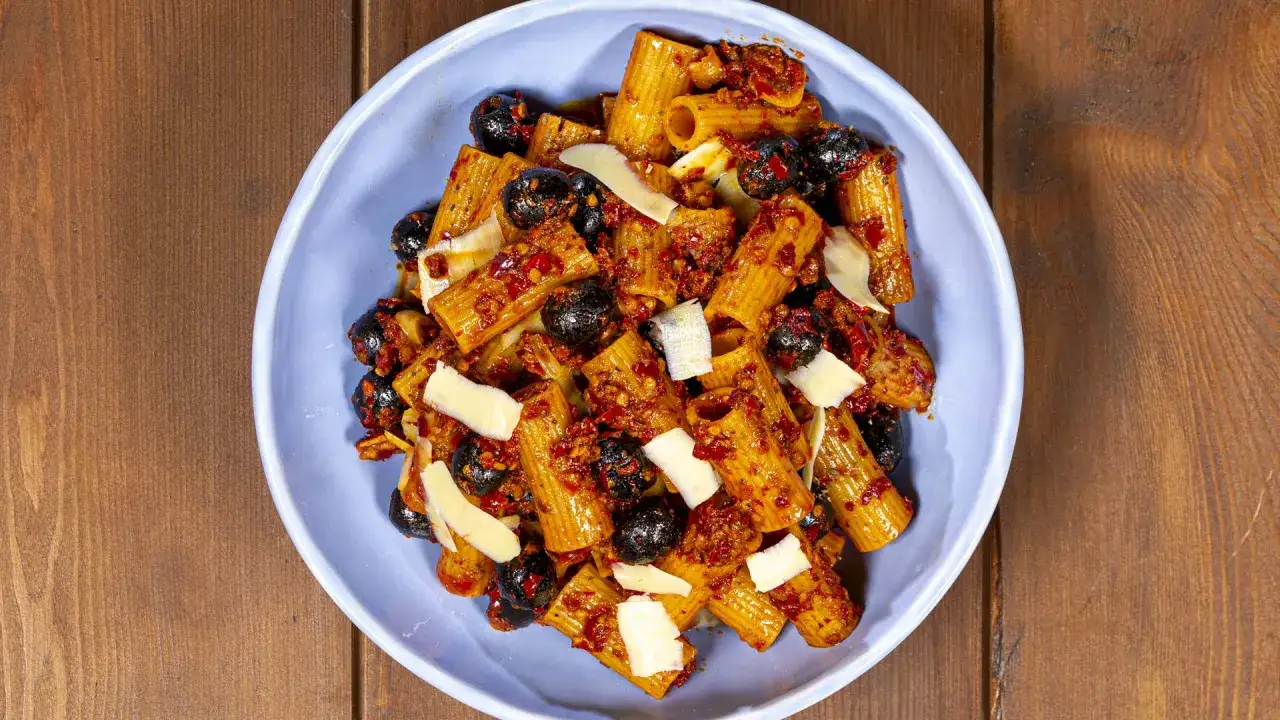 Rigatoni con nduja, olive nere e scaglie di parmigiano in una ciotola blu su sfondo di legno.
