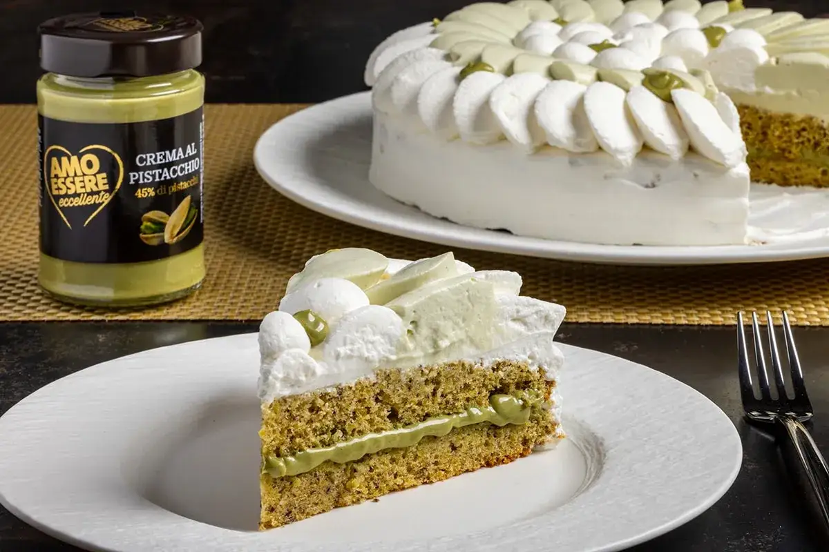 Una fetta di torta soffice con crema al pistacchio, perfetta per una ricetta golosa. Accanto, un barattolo di crema al pistacchio.