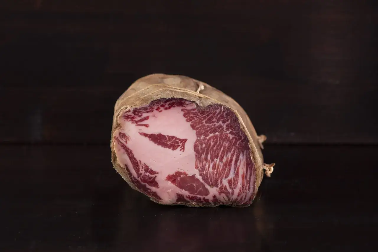 Un pezzo di coppa stagionata, con la sua carne rossa marmorizzata e il grasso bianco, avvolto in tela.