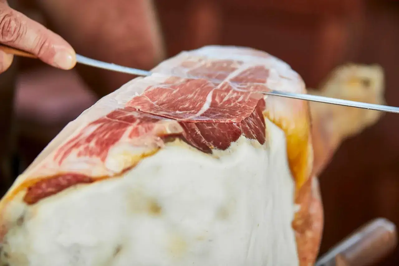 Mano esperta che affetta sottili fette di prosciutto crudo, mostrando la tecnica per come tagliare il prosciutto crudo.