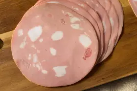 Fette di mortadella su tagliere di legno. Scopri come si fa la mortadella, un salume rosa con cubetti di grasso bianco.