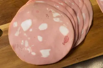 Fette di mortadella su tagliere di legno. Scopri come si fa la mortadella, un salume rosa con cubetti di grasso bianco.