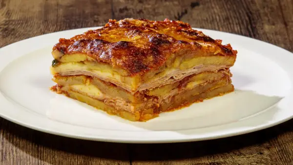 Una fetta di parmigiana di patate, stratificata con formaggio e salumi, servita su un piatto bianco.