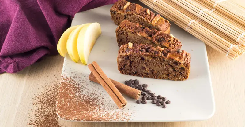 Fette di plumcake mela e cannella su piatto bianco, con mele, cannella, gocce di cioccolato e cacao in polvere.