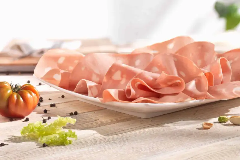Fette di mortadella su piatto bianco, con pomodoro, lattuga e pepe. La differenza tra mortadella e bologna è nella qualità e negli ingredienti.