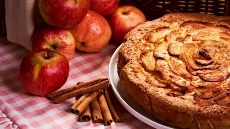 Torta mele, noci e cannella dorata, con mele fresche e bastoncini di cannella su una tovaglia a quadri rosa.